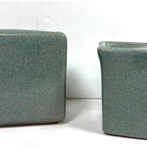 May include: Two ceramic containers with a speckled teal glaze. The object on the left is a wide, rectangular box with rounded corners. The object on the right is a smaller, tapered square vessel with an open top and a slightly flared rim.