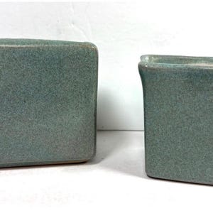 May include: A rectangular ceramic container with a smooth, matte, sage green finish positioned next to a smaller, square-shaped ceramic vessel featuring the same sage green glaze and a slightly flared top rim, both displaying a consistent texture.