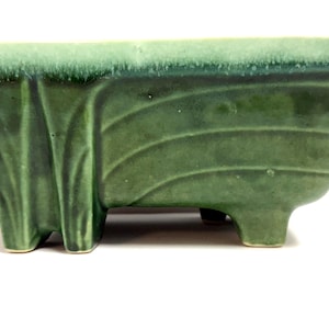 May include: A rectangular, green ceramic planter with a textured, ridged design. The planter has a slightly flared shape and is supported by four small feet. The top edge has a lighter green color.