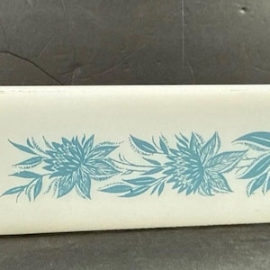 May include: A rectangular white glass loaf pan featuring a repeating blue floral pattern along the center of the side. The dish has a smooth, slightly tapered shape with a wide rim, constructed from opaque white milk glass with blue ink detailing.
