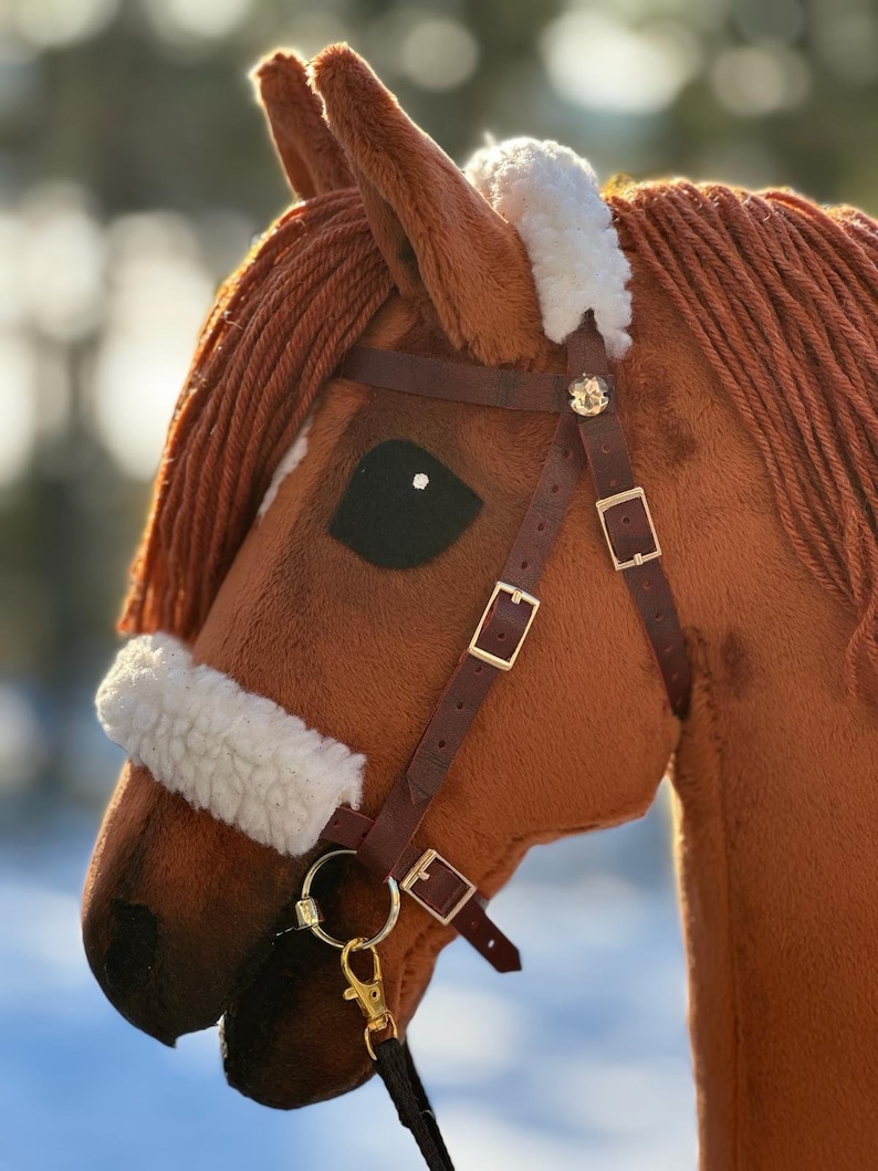 Pu&ograve; includere: Una testa di cavallo di peluche marrone con una fascia frontale in pile bianco e una briglia in pelle marrone con una fibbia dorata e un anello dorato. Il cavallo ha un occhio nero e un naso nero.