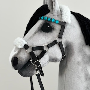Puede incluir: Una cabeza de caballo gris con una crin negra y un cabezada de cuero negro con una banda de frente de diamantes de imitaci&oacute;n turquesa. La cabezada tiene una rienda de cuero negro.