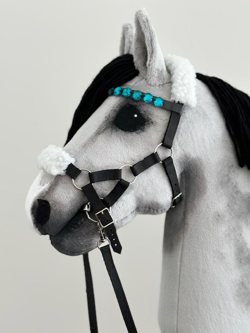 Puede incluir: Una cabeza de caballo gris con una crin negra y un cabezada de cuero negro con una banda de frente de diamantes de imitaci&oacute;n turquesa. La cabezada tiene una rienda de cuero negro.
