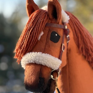 Pu&ograve; includere: Una testa di cavallo di peluche marrone con una fascia frontale in pile bianco e una briglia in pelle marrone. Il cavallo ha un occhio nero e un naso nero.