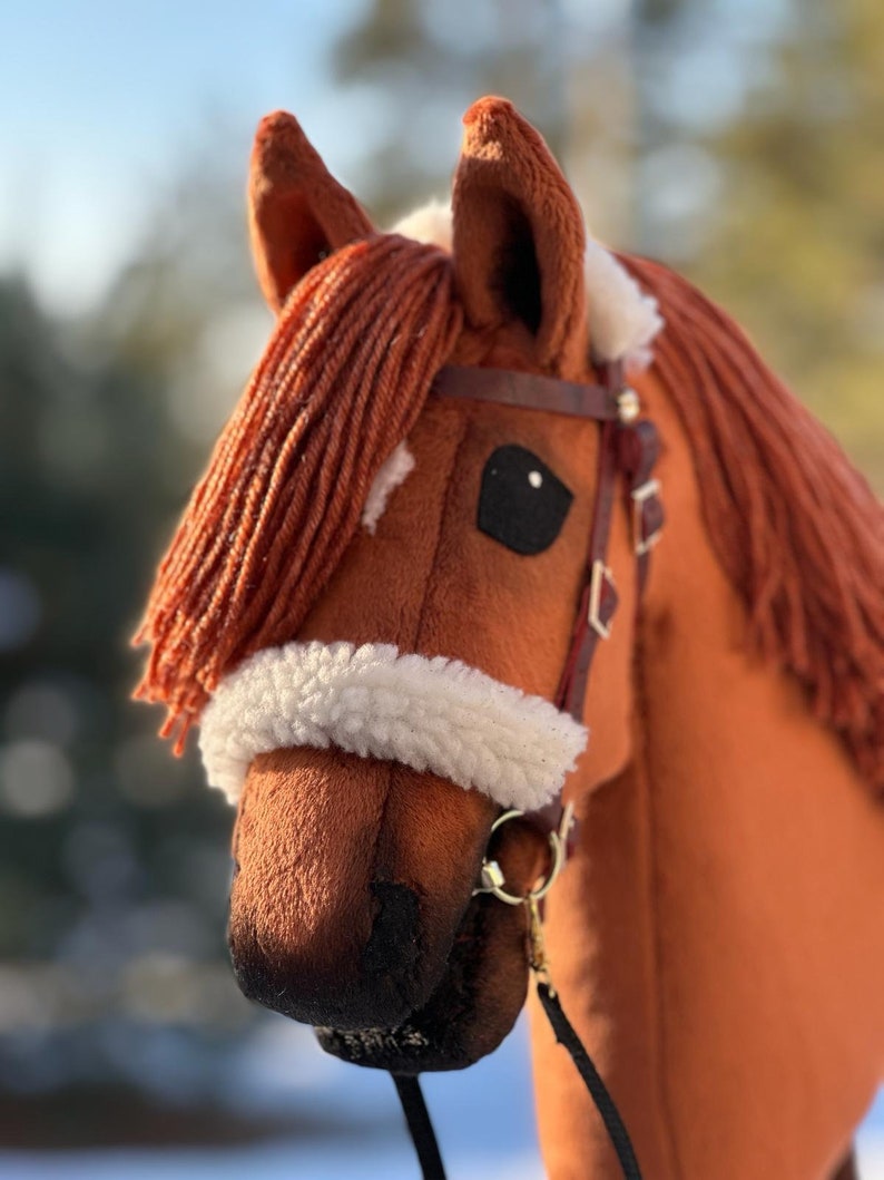 Pu&ograve; includere: Una testa di cavallo di peluche marrone con una fascia frontale in pile bianco e una briglia in pelle marrone. Il cavallo ha un occhio nero e un naso nero.