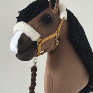 Peut inclure: Un cheval à bascule marron avec une crinière noire et un licol blanc et beige. Le cheval jouet a une corde tressée marron attachée au licol. Le cheval est en tissu doux et est conçu pour les enfants.