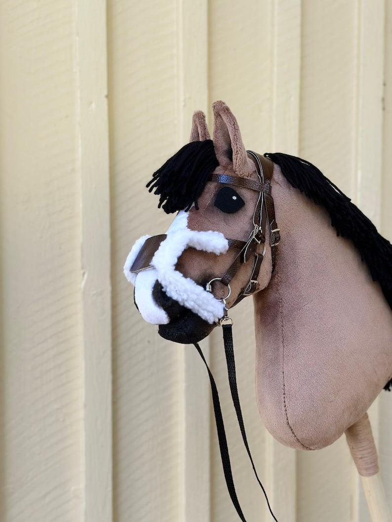Peut inclure: Une t&ecirc;te de cheval en peluche marron et blanche avec une crini&egrave;re et une queue noires. Le cheval porte une bride en cuir marron avec un nez en polaire noir et une longe noire.