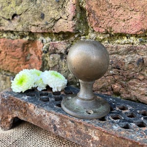 May include: An antique brass door knob with a spherical handle and a circular base. The knob is positioned against a weathered brick wall and a rusty metal grate. Two white and green flowers are in the foreground.