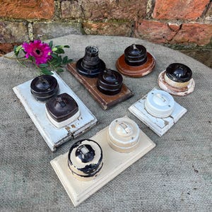 May include: A selection of antique light switches in diverse designs and colours, including black, brown, white, and cream. Some are mounted on wooden or painted bases, displaying a variety of styles and finishes.