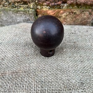 Może przedstawiać: Ciemnobrązowa, kulista gałka z kr&oacute;tką, cylindryczną podstawą. Gałka jest umieszczona na teksturowanej tkaninie o neutralnym odcieniu, z zwietrzałą ceglaną ścianą w tle. Gałka wydaje się być wykonana z drewna.