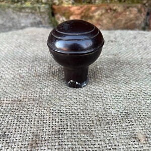 Może przedstawiać: Ciemnobrązowy uchwyt do szafki w stylu vintage z okrągłym, rowkowanym wierzchem. Uchwyt ma kr&oacute;tki trzon i jest umieszczony na tle teksturowanej, neutralnej kolorystycznie tkaniny. Konstrukcja uchwytu sugeruje, że jest przeznaczony do renowacji mebli.