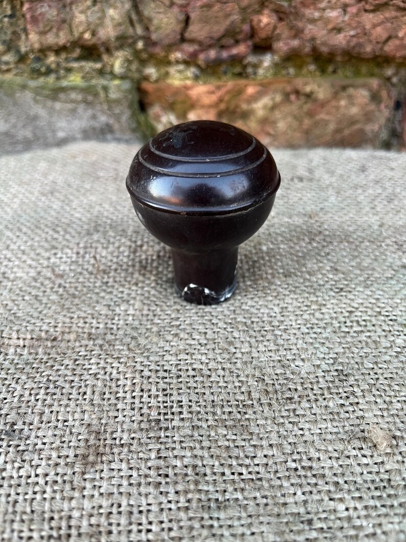 Może przedstawiać: Ciemnobrązowy uchwyt do szafki w stylu vintage z okrągłym, rowkowanym wierzchem. Uchwyt ma kr&oacute;tki trzon i jest umieszczony na tle teksturowanej, neutralnej kolorystycznie tkaniny. Konstrukcja uchwytu sugeruje, że jest przeznaczony do renowacji mebli.