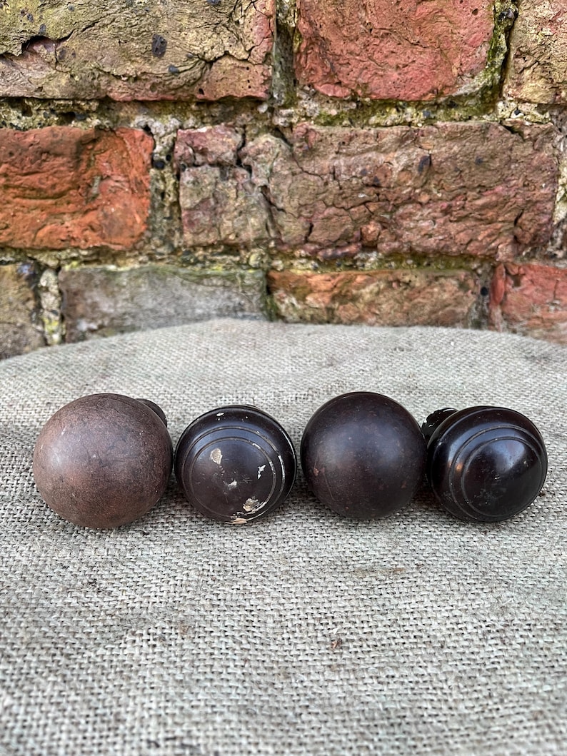 Może przedstawiać: Cztery antyczne drewniane gałki do drzwi w r&oacute;żnych odcieniach brązu. Gałki są okrągłe z gładkimi lub rowkowanymi wzorami. Ułożone są na teksturowanej, neutralnej powierzchni, z ceglaną ścianą w tle.