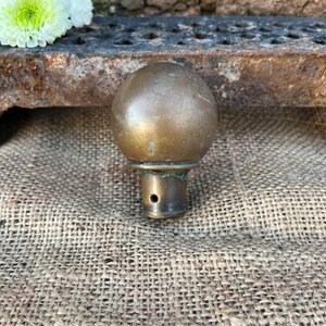 May include: A vintage brass knob with a rounded top and a cylindrical base. The knob has a weathered, aged appearance, with visible patina and slight discoloration. The background includes a textured hessian surface and a metal grate.