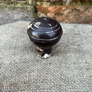 Może przedstawiać: Ciemnobrązowa gałka do drzwi w stylu antycznym z zaokrągloną g&oacute;rą i cylindryczną podstawą. Gałka posiada trzy srebrne paski wok&oacute;ł g&oacute;ry. Podstawa ma odpryski białej farby. Gałka jest umieszczona na teksturowanej, szarej tkaninie.