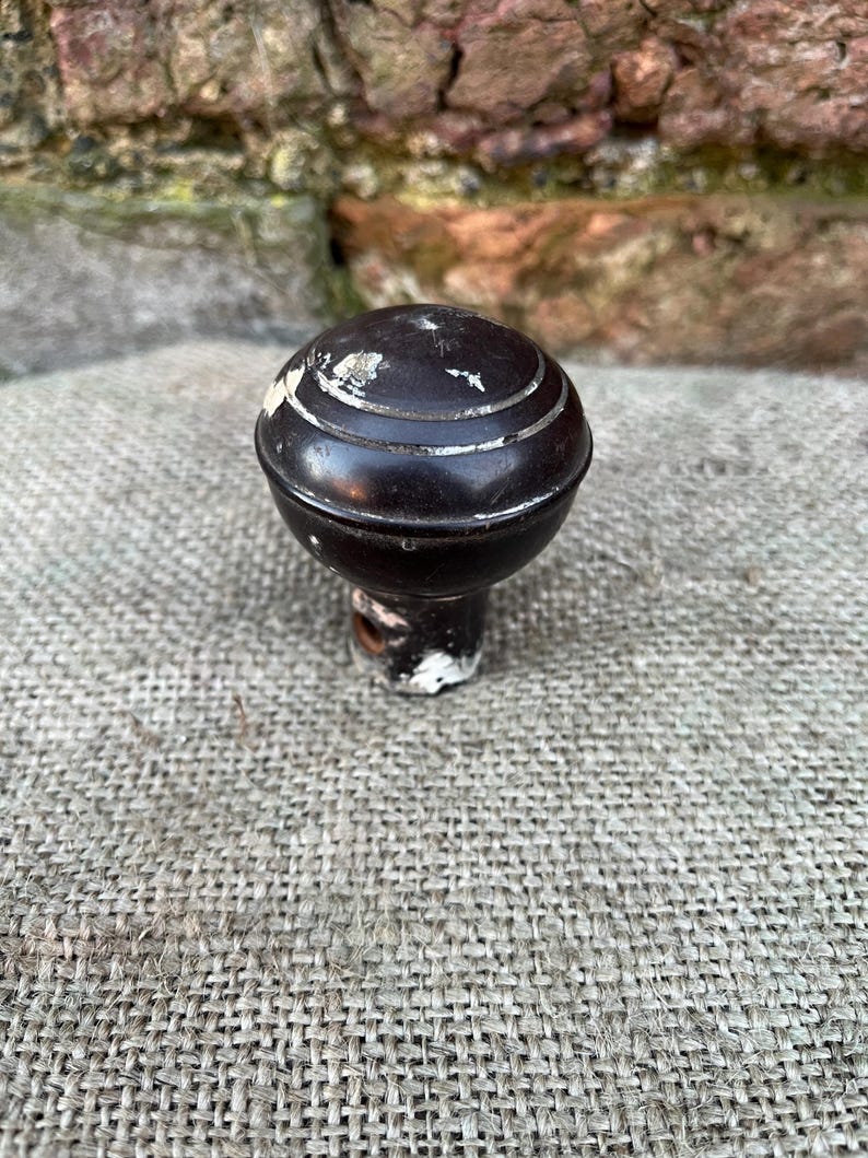 Może przedstawiać: Ciemnobrązowa gałka do drzwi w stylu antycznym z zaokrągloną g&oacute;rą i cylindryczną podstawą. Gałka posiada trzy srebrne paski wok&oacute;ł g&oacute;ry. Podstawa ma odpryski białej farby. Gałka jest umieszczona na teksturowanej, szarej tkaninie.