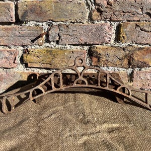 May include: A decorative, rusty metal hanger with an ornate design. The hanger is curved with cut-out patterns and a circular top. The background shows a weathered brick wall and burlap fabric.