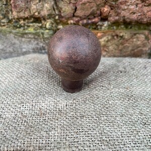 Może przedstawiać: Ciemnobrązowy, kulisty uchwyt do szafki z kr&oacute;tkim, cylindrycznym trzpieniem. Gałka ma teksturowany, postarzany wygląd, sugerujący, że jest wykonana z metalu lub podobnego materiału. Gałka jest umieszczona na neutralnym, teksturowanym tle.