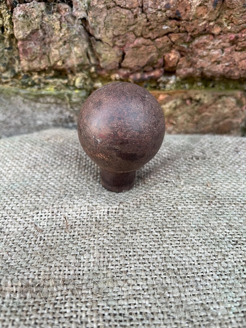 Może przedstawiać: Ciemnobrązowy, kulisty uchwyt do szafki z kr&oacute;tkim, cylindrycznym trzpieniem. Gałka ma teksturowany, postarzany wygląd, sugerujący, że jest wykonana z metalu lub podobnego materiału. Gałka jest umieszczona na neutralnym, teksturowanym tle.