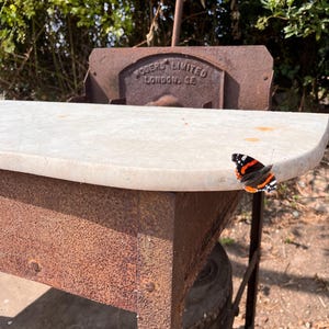 May include: A vintage metal table with a marble top. The table has a rusty finish and a butterfly with orange, black, and white wings is perched on the edge. The text "ROSERS LIMITED LONDON. GE" is visible.
