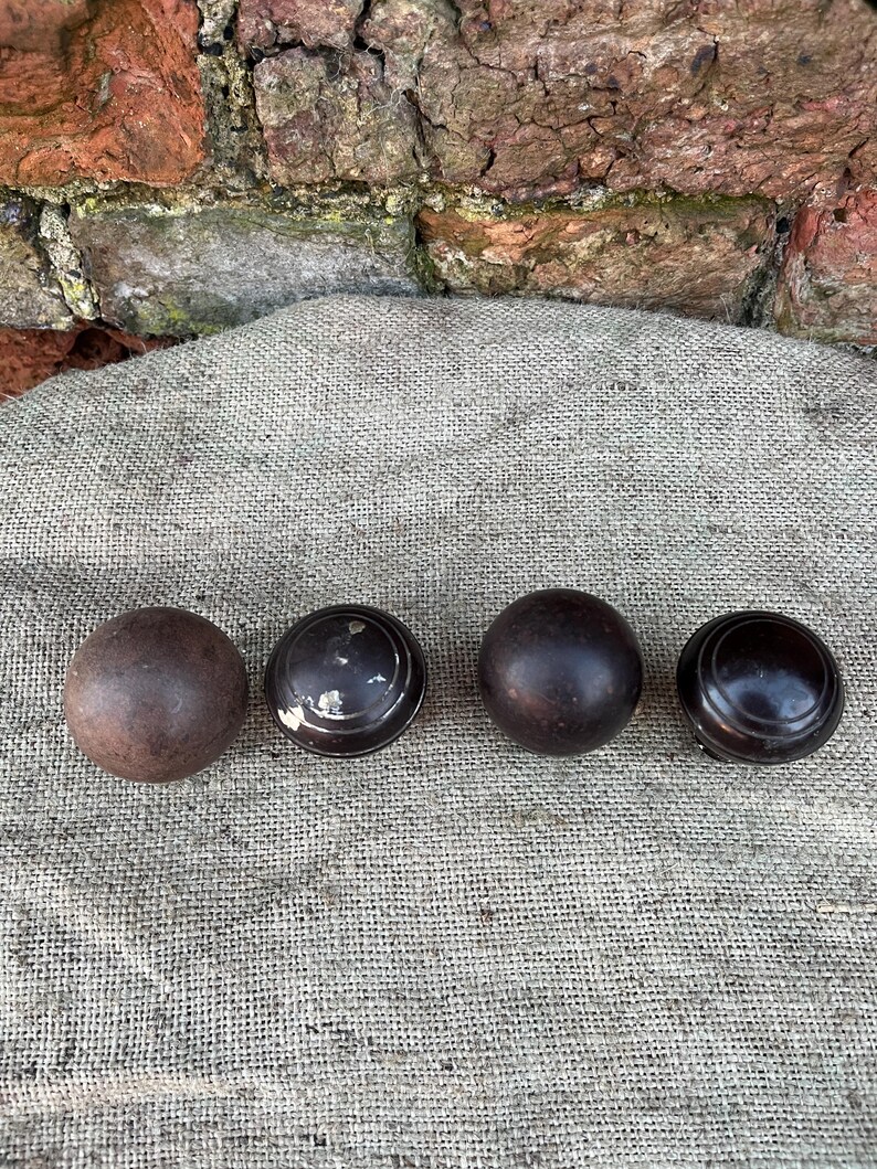 Może przedstawiać: Cztery antyczne drewniane gałki w r&oacute;żnych odcieniach brązu. Gałki ułożone są w rzędzie na teksturowanej, neutralnej powierzchni. Gałki mają okrągły kształt, niekt&oacute;re z płaskim, tarczowatym wzorem.