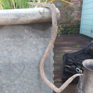 May include: A weathered, rusty metal tool with a curved design and a pointed end. It is hanging over the edge of a grey metal container. The tool's shape suggests it could be a hook or a lever, possibly for gardening.