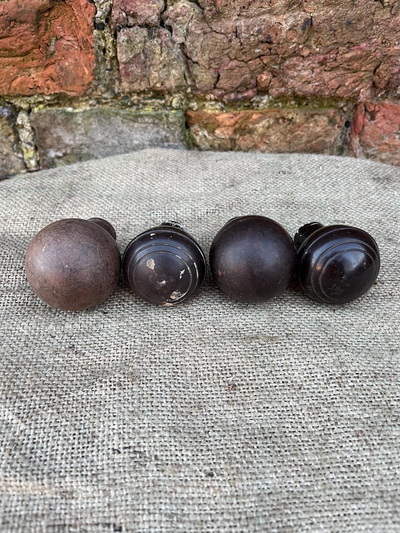 Może przedstawiać: Cztery antyczne drewniane gałki w r&oacute;żnych kształtach i odcieniach brązu. Gałki ułożone są na teksturowanej, neutralnej powierzchni, z tłem z wyblakłej cegły.