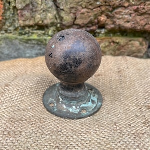 May include: Weathered bronze door knob with a spherical top and a circular base. The knob has a worn appearance with dark brown and black tones, and a hint of green patina on the base. The knob is set against a hessian surface.