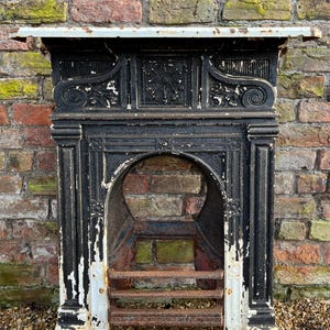 May include: An antique black cast iron fireplace mantel with white detailing. The mantelpiece features ornate scrollwork and a decorative central panel. The fireplace has a curved opening and a rusted grate. It is set against a weathered brick wall.