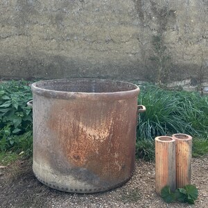 May include: A large, weathered metal container with a cylindrical shape and two small handles. The container has a rusted, aged appearance. Two smaller, cylindrical objects with vertical grooves stand nearby.
