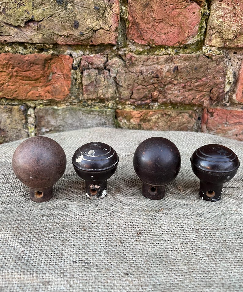 Może przedstawiać: Cztery antyczne drewniane gałki w r&oacute;żnych odcieniach brązu. Gałki mają okrągły kształt z ozdobną podstawą i otworem do montażu. Ułożone są na tkaninie w neutralnym kolorze.