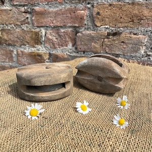 May include: Two weathered wooden pulley blocks with central metal pins, one cylindrical and one oval-shaped, arranged alongside three small white daisies with yellow centres on a textured brown fabric surface.