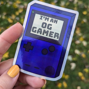May include: A blue and silver holographic sticker with a retro game console design. The sticker says "I'm an OG Gamer" in white text.