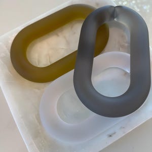 May include: Three oval-shaped chain links with a smooth, matte finish. One link is olive green, one is charcoal grey, and one is translucent white. The links are arranged in an overlapping pattern, showcasing their rounded, continuous loop design.
