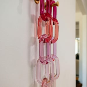 以下が含まれることがあります： チェーンリンクデザインの装飾壁掛けフック。リンクは透明なピンクと赤のガラス製で、白い壁に取り付けられた金色の金属フックから吊り下げられています。リンクはピンクと赤のさまざまな色合いです。