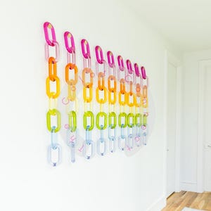 May include: A colorful wall art piece featuring a series of acrylic chain links in a rainbow of colors. The chains are mounted on a white wall, creating a vibrant and modern decorative element. The links are in shades of pink, orange, yellow, green, and blue.