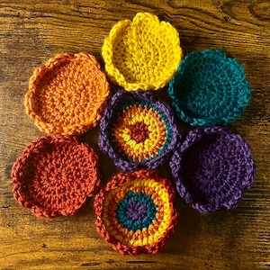 May include: A set of seven circular crochet coasters featuring scalloped edges. The collection includes solid yellow, orange, teal, and purple coasters, alongside three multicoloured coasters with concentric rings of yellow, orange, red, and blue.