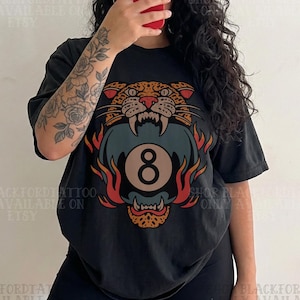Może przedstawiać: Czarny t-shirt z motywem głowy tygrysa i grafiką kuli 8 w kolorach pomarańczowym, czerwonym i niebieskim. Tekst "SHOP BLACKFORDTATTOO ONLY AVAILABLE ON ETSY" jest wydrukowany na górze i na dole obrazu. Widoczny tatuaż na ramieniu.