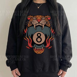 May include: Black sweatshirt featuring a colorful graphic of a snarling leopard head with an eight ball in its mouth, surrounded by flames. The text "SHOP BLACKFORDTATTOO ONLY AVAILABLE ON ETSY" is printed around the image.
