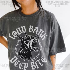 Pode incluir: Camiseta cinza escuro com um desenho gráfico em preto e branco de uma cabeça de cachorro rosnando dentro de um círculo de arame farpado. O texto "LOUD BARK DEEP BITE" está impresso acima e abaixo. A camiseta tem um visual vintage.