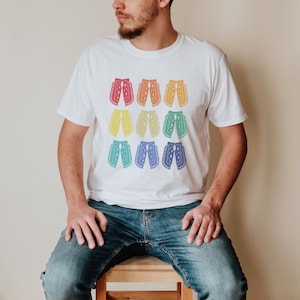 Puede incluir: Una camiseta blanca con un patrón arcoíris de nueve pares de pantalones de vaquero. Los pantalones están delineados en negro y tienen estrellas blancas.