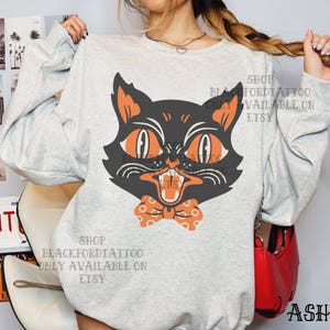 May include: A light gray sweatshirt featuring a black cat graphic with orange eyes and a bow tie. The cat has an open mouth and a slightly angry expression. The text "SHOP BLACKFORDTATTOO ONLY AVAILABLE ON ETSY" is printed on the sweatshirt.