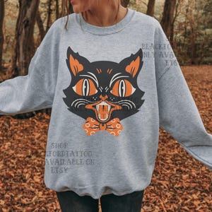 May include: A light gray sweatshirt featuring a black cat graphic with orange eyes, ears, and a bow tie. The cat has an open mouth with visible teeth. The text "BLACKFORDTATTOO ONLY AVAILABLE ON ETSY" is printed on the sweatshirt.
