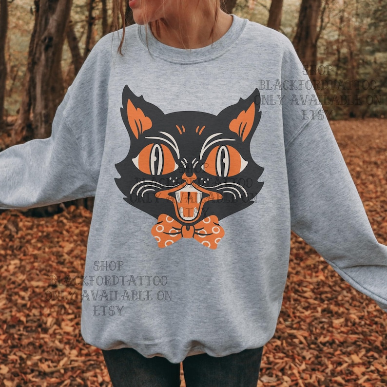 May include: A light gray sweatshirt featuring a black cat graphic with orange eyes, ears, and a bow tie. The cat has an open mouth with visible teeth. The text "BLACKFORDTATTOO ONLY AVAILABLE ON ETSY" is printed on the sweatshirt.