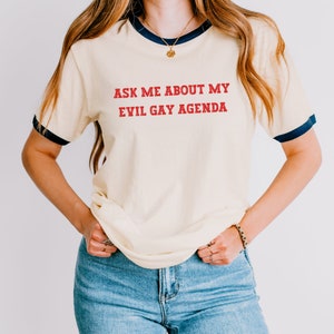 May include: Cream-colored ringer t-shirt with navy blue trim and the text "ASK ME ABOUT MY EVIL GAY AGENDA" in red. The shirt is paired with blue jeans. The overall style is casual and comfortable.