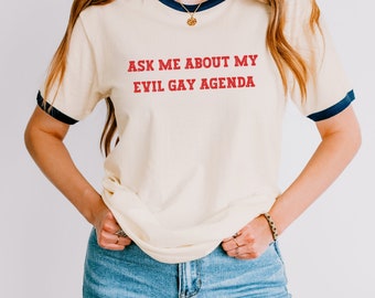 Könnte beinhalten: Cremefarbenes Ringer-T-Shirt mit marineblauen Abschlüssen und dem roten Text "ASK ME ABOUT MY EVIL GAY AGENDA". Das Shirt wird mit einer blauen Jeans kombiniert. Der Stil ist lässig und bequem.