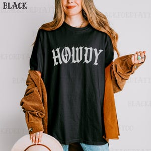 May include: A black t-shirt with the word "Howdy" printed in white on the front. The shirt is being worn by a person with long brown hair.