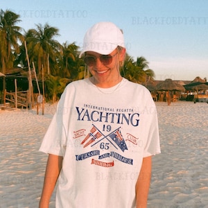 Op de afbeelding: Wit T-shirt met een grafisch ontwerp met de tekst "YACHTING" met gekruiste vlaggen en de tekst "International Regatta 19 65 Classic and Vintage Sailing Yacht Club".