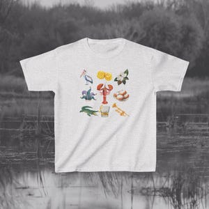 May include: A light gray t-shirt with a colorful illustration of various symbols associated with New Orleans, including a fleur-de-lis, a crawfish, a trumpet, a glass of whiskey, and a magnolia flower.