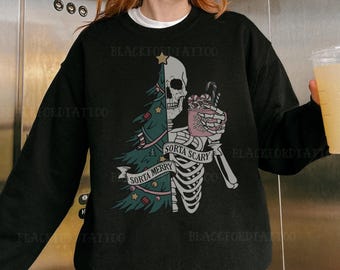 Op de afbeelding: Zwart sweatshirt met een skelet dat een mok warme chocolademelk vasthoudt. Het skelet staat voor een kerstboom met de tekst "Sorta Merry" en "Sorta Scary" aan weerszijden van de boom.